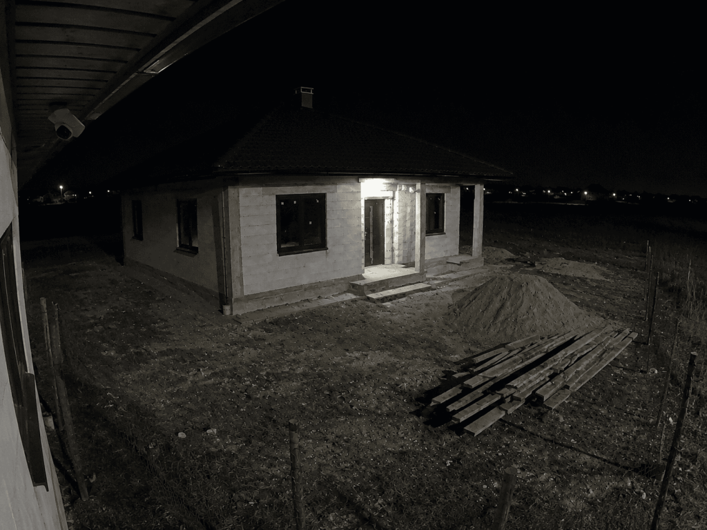 Nocny widok z kamery monitoringu na plac budowy domu, wejście do budynku, piasek i deski.